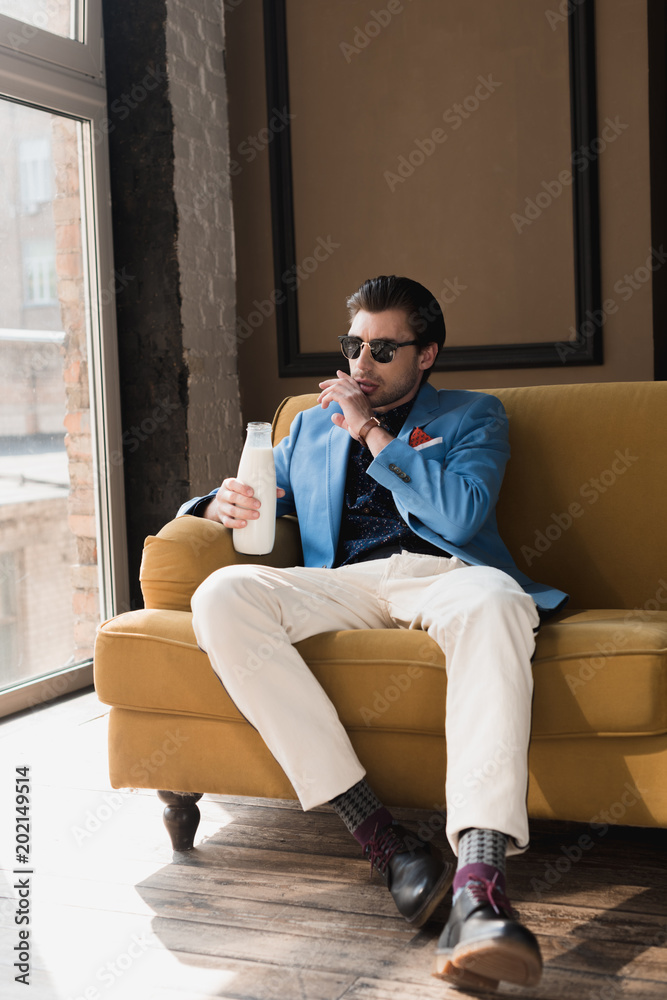 attractive young man in stylish suit sitting on couch with bottle of milk and wiping mouth with hand