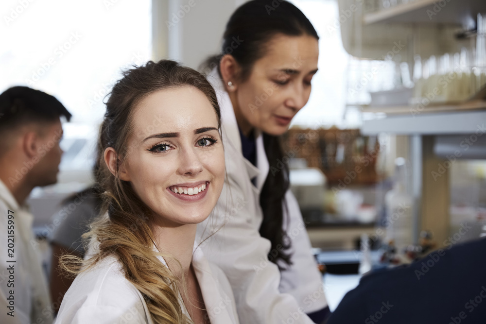 Portrait of smiling young female student sitting against teacher friends in chemistry laboratory Portrait of smiling young female student sitting against teacher friends in chemistry laboratory