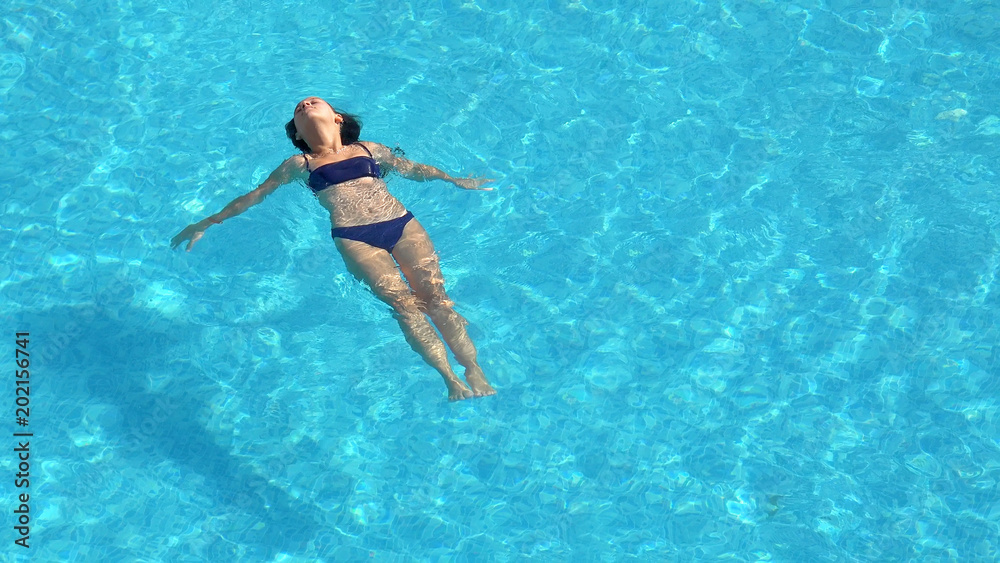 Young woman swimming in the crystal-clear pool laying on water surface