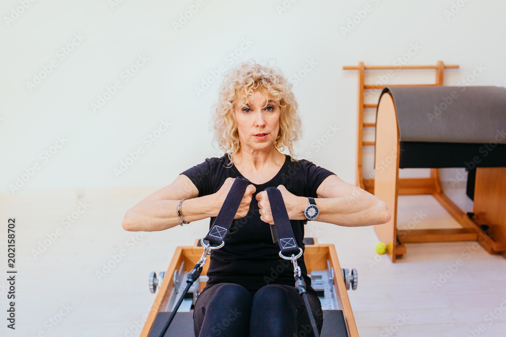 Pilates reformer eldery blond curly woman gym fitness teacher workout back  and hands exercise on pilates reformer device - pilates, fitness, sport,  training and people concept. Stock Photo