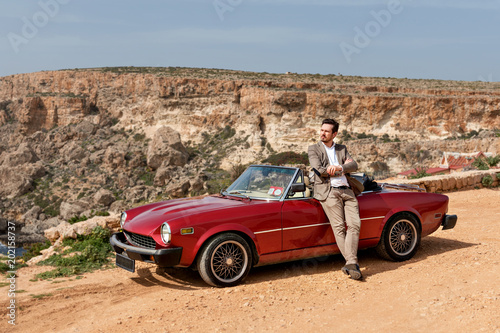 an attractive handsome man mate is leaning on a retro car in a classic suit on a sunny afternoon on a sandy desert road