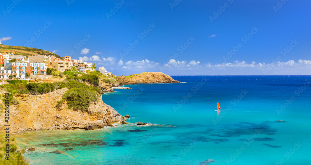Fototapeta premium Windsurf with red sail rides on sea on background rocky shore. View from sunny Livadi beach in resort village Bali. Rethymno, Crete Greece. Extreme water sports as active recreation on Sunny vacations