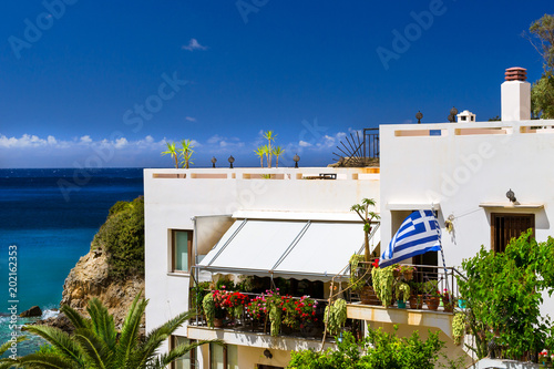 Modern and classic resort Greek architecture, white building stands on shore of Cretan sea. Greek flag is set on the facade of the building. Resort village Bali, Rethymno, Crete, Greece
