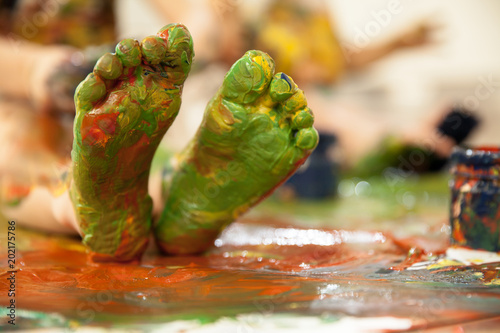  Foot feet are painted with colors. A child is sitting in the colors of gouache.