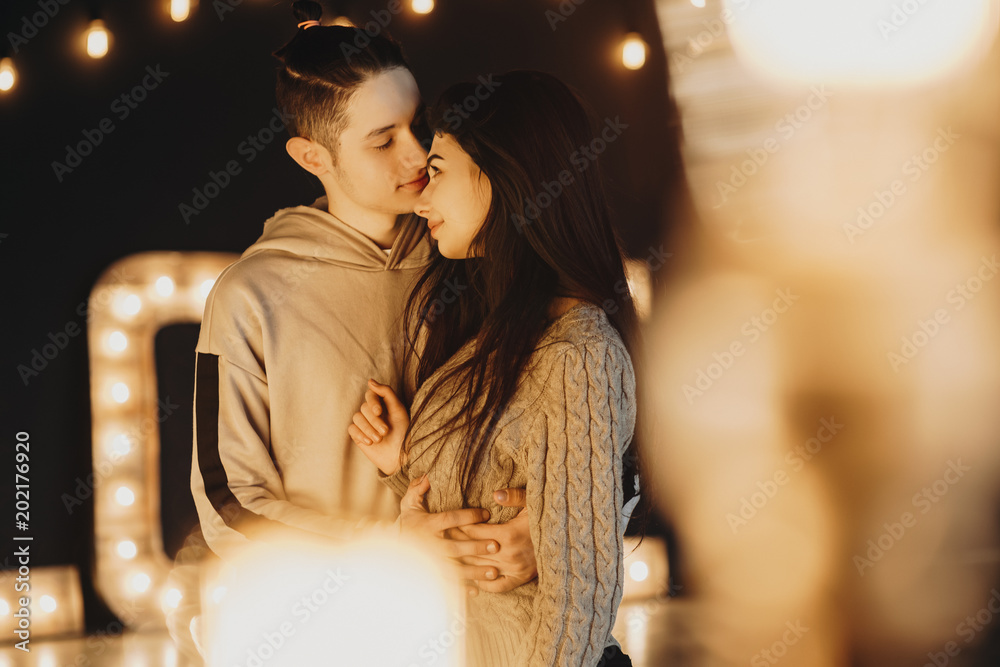 Young and beautiful caucasian couple embracing and and looking to each other with love through warm lights in a black room decorated with warm lights.