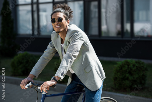 stylish young african american man in sunglasses sitting on bicycle and smiling at camera