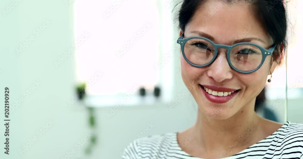 Smiling young Asian businesswoman wearing glasses standing with her arms crossed in a modern office with colleagues in the background Smiling young Asian businesswoman wearing glasses standing with her arms crossed in a modern office with colleagues in the background