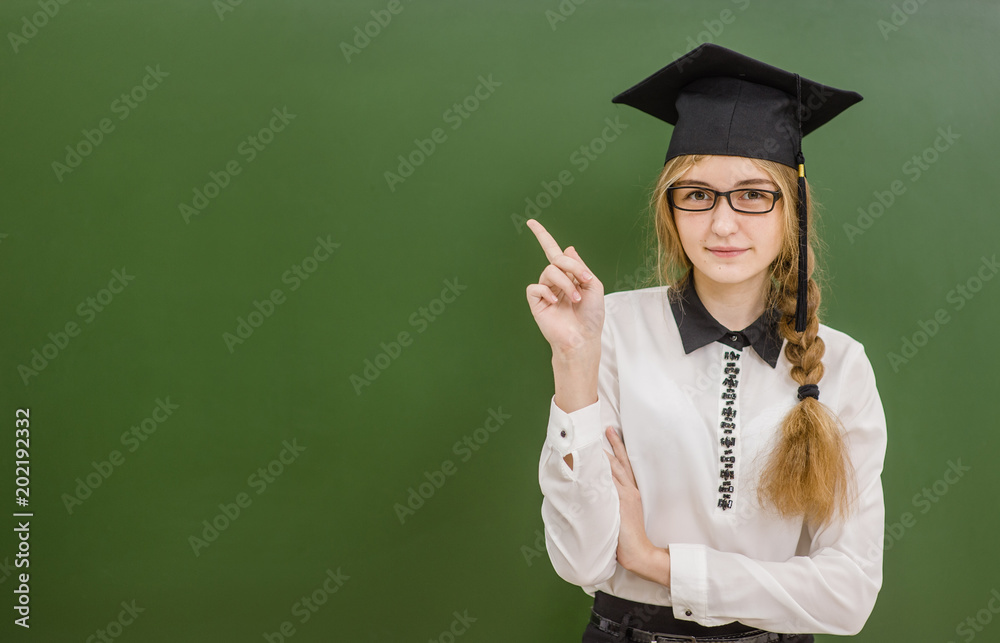 Teen girl in graduation cap standing near green chalkboard and pointing on empty space. Looking at camera Teen girl in graduation cap standing near green chalkboard and pointing on empty space. Looking at camera