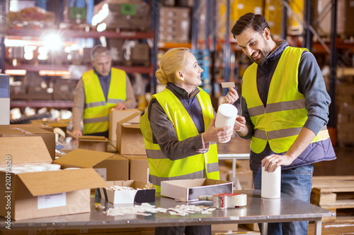 Wholesale production. Nice warehouse worker smiling and looking at each other while putting labels on the products 