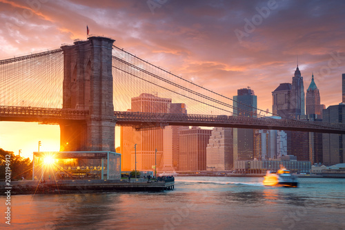 Fototapeta Naklejka Na Ścianę i Meble -  Famous Brooklyn Bridge in New York City with financial district - downtown Manhattan in background. Sightseeing boat on the East River and beautiful sunset over Jane's Carousel.