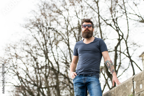 Barbershop and style concept. Man with beard on strict face, trees on background, defocused. Hipster with beard looks confident while standing outdoors. Bearded man wears modern sunglasses.