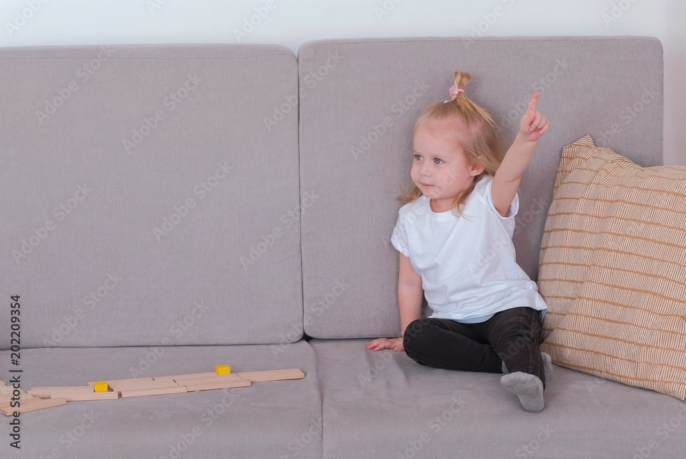 Naklejka premium Little charming girl sitting on the sofa talking and playing wood blocks.