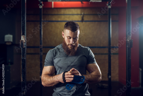 Фотография Portrait view of young bearded focused strong muscular shape bodybuilder man puts on wrist and hands support for exercises at the gym
