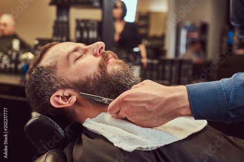 Professional hairdresser modeling beard with scissors and comb at the barbershop.