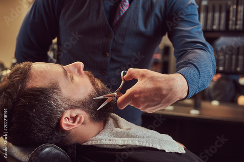 Professional hairdresser modeling beard with scissors and comb at the barbershop.