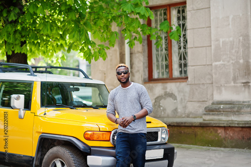 Stylish african american boy on gray sweater and black sunglasses posed on street against yellow car. Fashionable black business man. photo