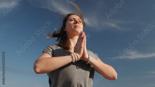 Young cheerful woman doing yoga asana on blue sky background. Girl in comfortable grey clothing meditating with namaste. Windy sunny weather. Health and grateful concept.