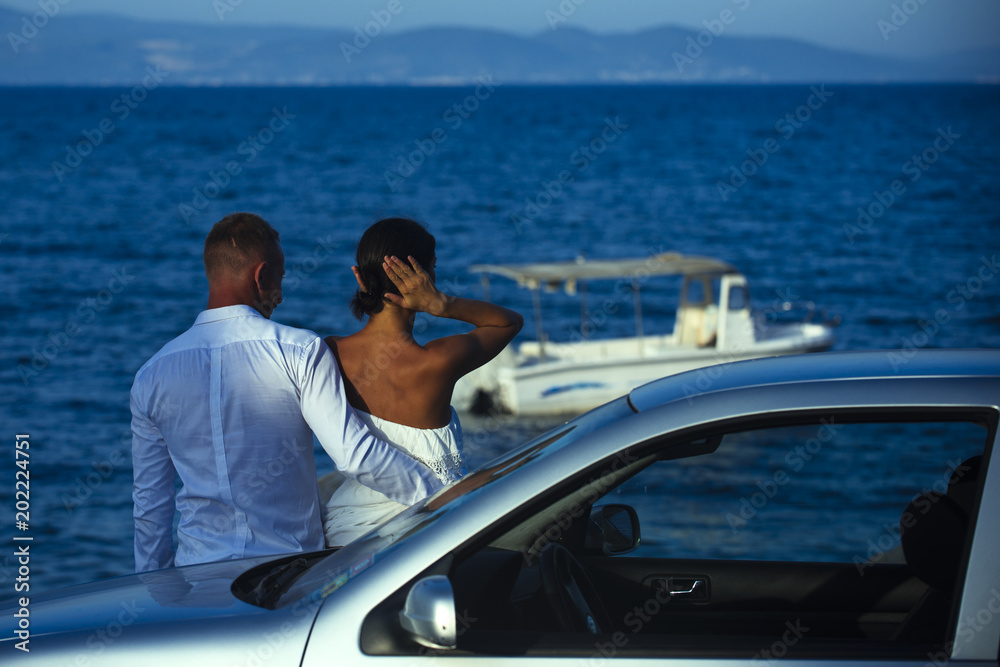 Fototapeta premium Couple in love stand near silver car, sea on background. Couple arrived on vacation, honeymoon. Man and woman looking at sea and boat, enjoy view. Romantic walk of couple near sea. Luxury rest concept