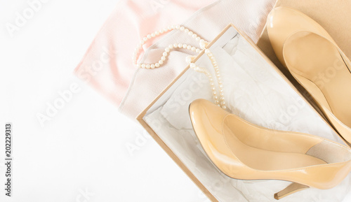 Cream heeled women's shoes, pink dress and pearls necklace on white background.