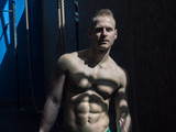 Attractive shirtless blond male bodybuilder in shorts indoors in dark gym, showing muscular torso and ripped abs