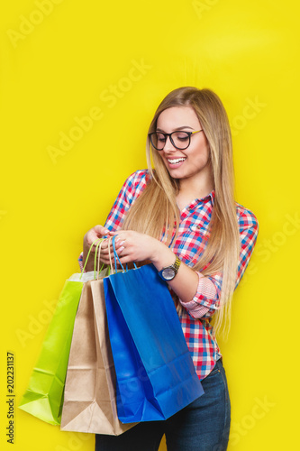 Photo of cheerful oung woman standing isolated over yellow wall background. Looking aside holding shopping bags.