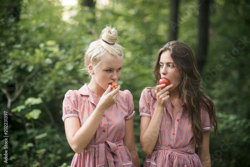 Camping in woods. Beautiful young girls wandering in forest. Two sisters in vintage dresses eating juicy tomatoes. Vitamins  nutrition and healthy diet concept