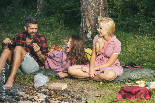 Happy friends camping in woods. Guy feeding his girfriend with fries sausages. Blond girl looking at her smiling friends photo