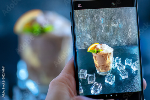 oman hand make shot of Orange Soda Creamsicle Ice Cream Float in glass decorated with piece of orange, straws and leaves on mint, surrounded by ice cubes on stone table backdrop by mobile phone photo