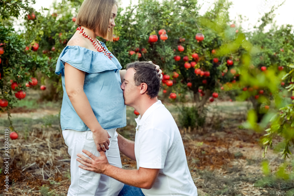 Portrait of happy family in the pomegrate fruit garden. Husband kneeling and kissing stomach of his pregnant wife. Fertility concept. Sunset light. soft selective focus, space for text. Portrait of happy family in the pomegrate fruit garden. Husband kneeling and kissing stomach of his pregnant wife. Fertility concept. Sunset light. soft selective focus, space for text.