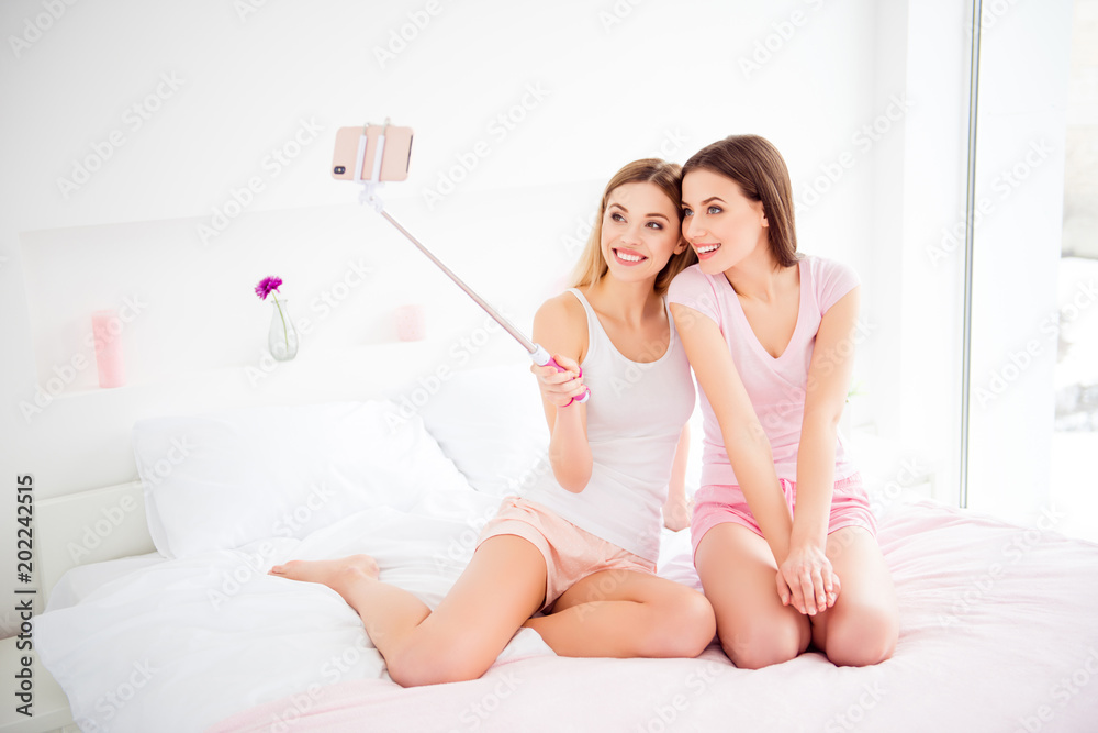 Portrait of pretty, charming, cheerful, cute, attractive, joyful girls sitting on bed in house, room, indoor, shooting selfie on front camera, using selfish stick and smart phone, spending free time