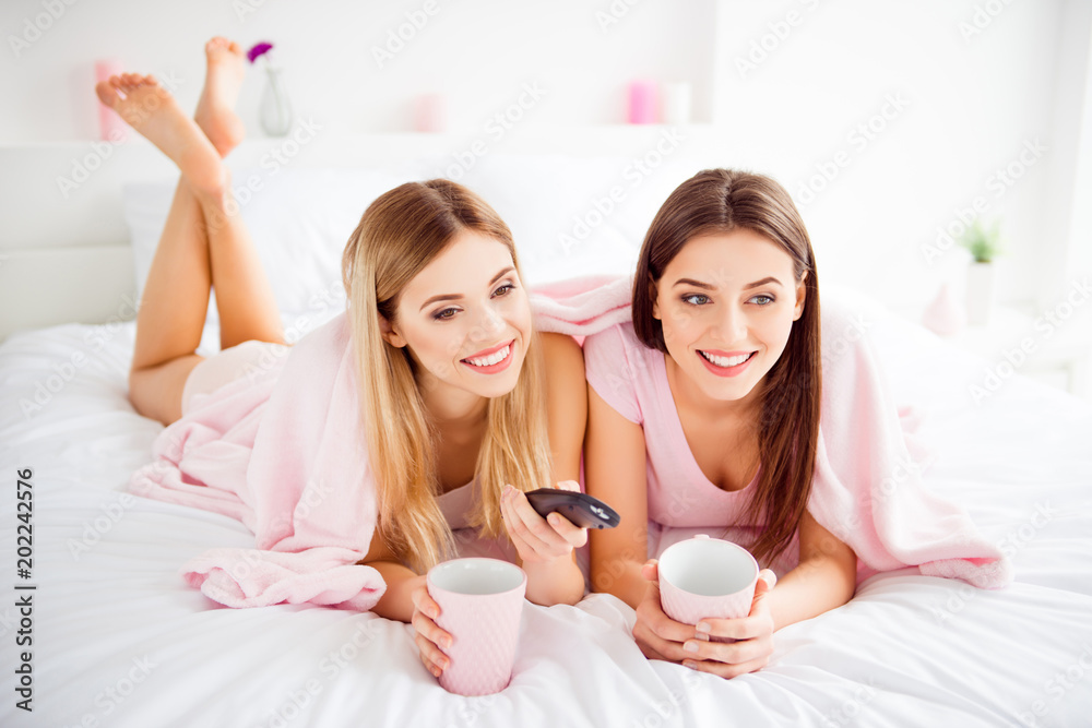 Pretty, charming, cute, cheerful, attractive girls in white and pink outfit having mugs of tea in hands, trying to find interesting program on tv with console, lying on stomach under coverlet