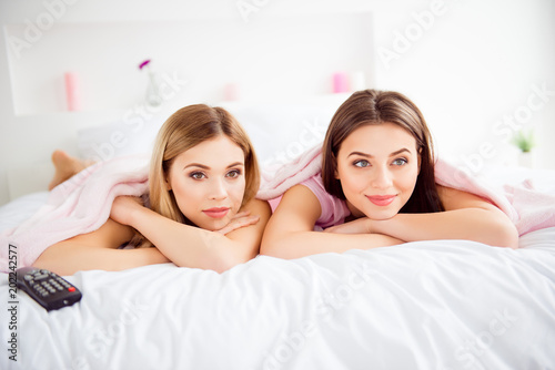 Portrait of pretty, charming, cute, cheerful, attractive, confident, successful girls in white and pink outfit, lying on stomach under coverlet, putting head, chin on folded, crossed arms, watching tv