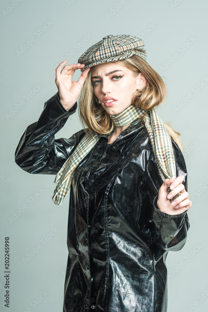 Fototapeta premium Girl with long hair and make up wears black cloak, plaid kepi, scarf, light background. Fashion accessories concept. Woman on calm face in cloak with accessories. Sexy lady in spring, autumn outfit.