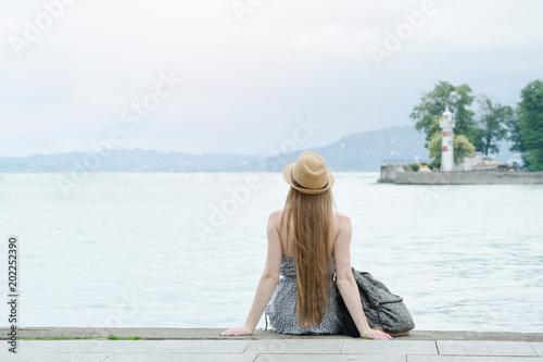 Girl in the hat sitting on the dock. Sea and small lighthouse in the distance. Back view © somemeans