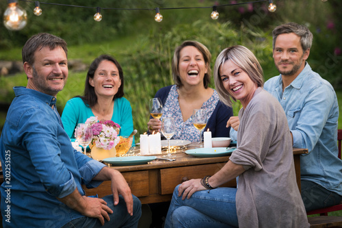 A summer evening of friends in their 40s gather around a table in the garden to share a meal and have fun together.