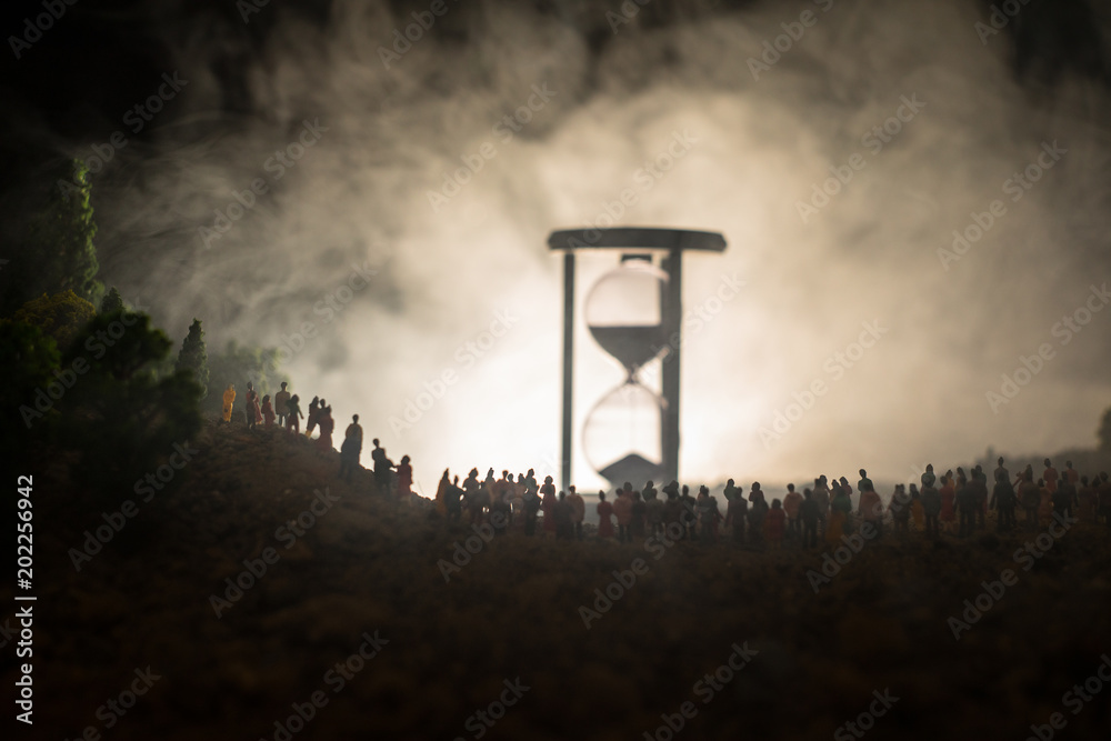 Silhouette of a large crowd of people in forest at night standing against a big hourglass with toned light beams on foggy background. Time concept.