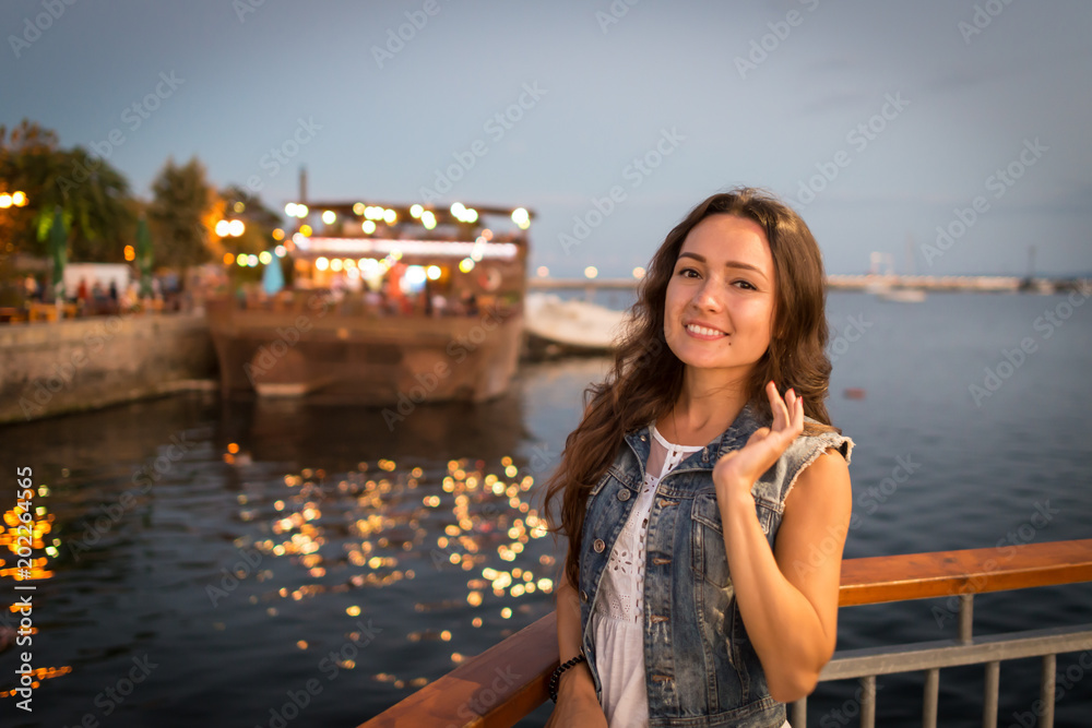 Pretty young woman walking on city promenade near sea in the evening. Soft colors. Street lights rays falls on her face. Happy girl enjoys vacation
