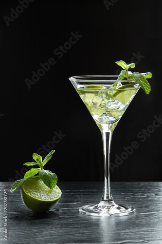 Summer fresh cocktail with lime slices, crushed ice and mint leaves in a martini glass backlit on black background