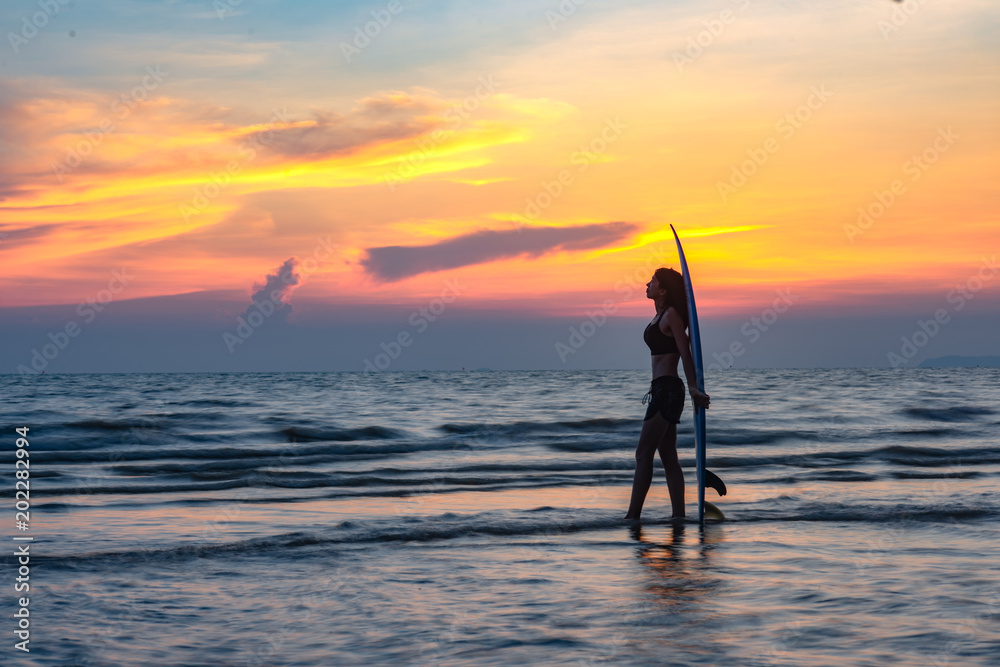 woman holding a surfboard in hand laying on back beside, on returning home station after played surf in the sea at sunset