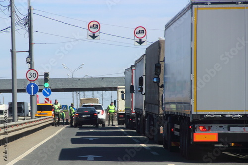 The queue of trucks with semi-trailers to the control point in the spring against the blue sky, inspectors in yellow jackets and camouflage check the documents of the driver of the car