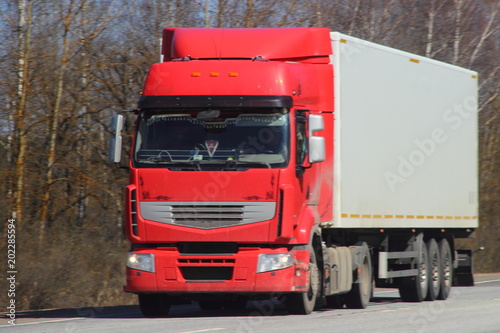 Transportation logistics - red truck with white semi-trailer on asphalt road in spring against blue sky forest, front view