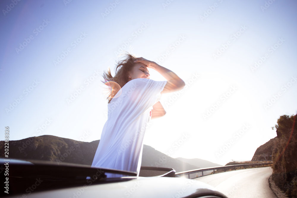 Happy excited beautiful young woman, free and careless teenager living the american dream on sunny empty road, stands up in convertible cabriolet car, wind in her hair, she laughs and smiles