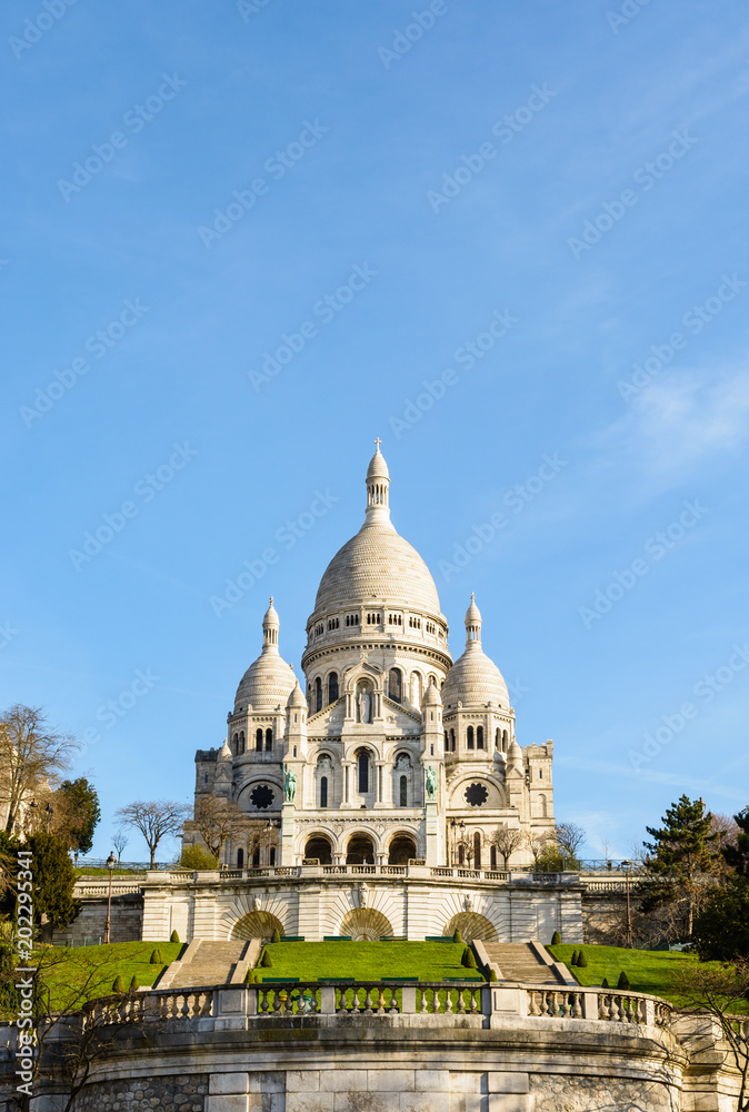 The basilica of the Sacred Heart of Paris under a blue sky in springtime seen from the bottom of the Louise Michel park, with the terrace and stairways on the foreground.
