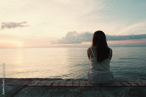Silhouette of young woman sitting alone on back side outdoor at tropical island beach missing boyfriend and family in summer sunset.