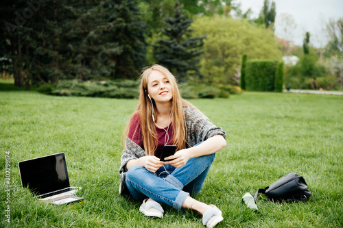 Smiling young blonde woman sitting on grass and listening music outside in park © Vulp