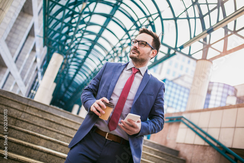 Businessman using mobile phone outside of office buildings in the background. Young caucasian man holding smartphone for business work and drink coffee to go.