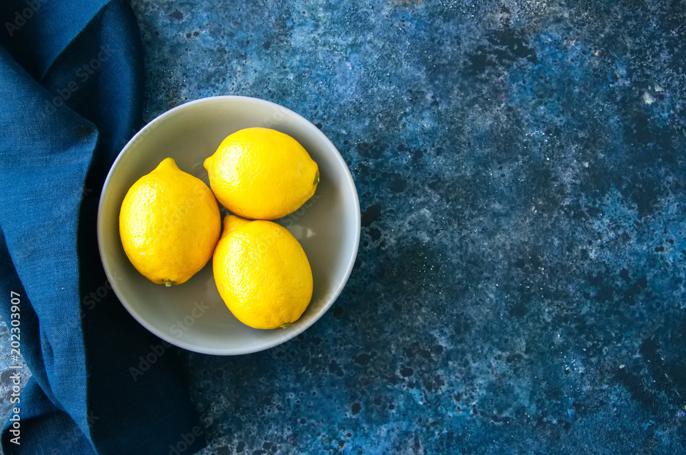 Three ripe yellow lemons in a grey bowl on a blue stone background. Top view and copy space. Three ripe yellow lemons in a grey bowl on a blue stone background. Top view and copy space.