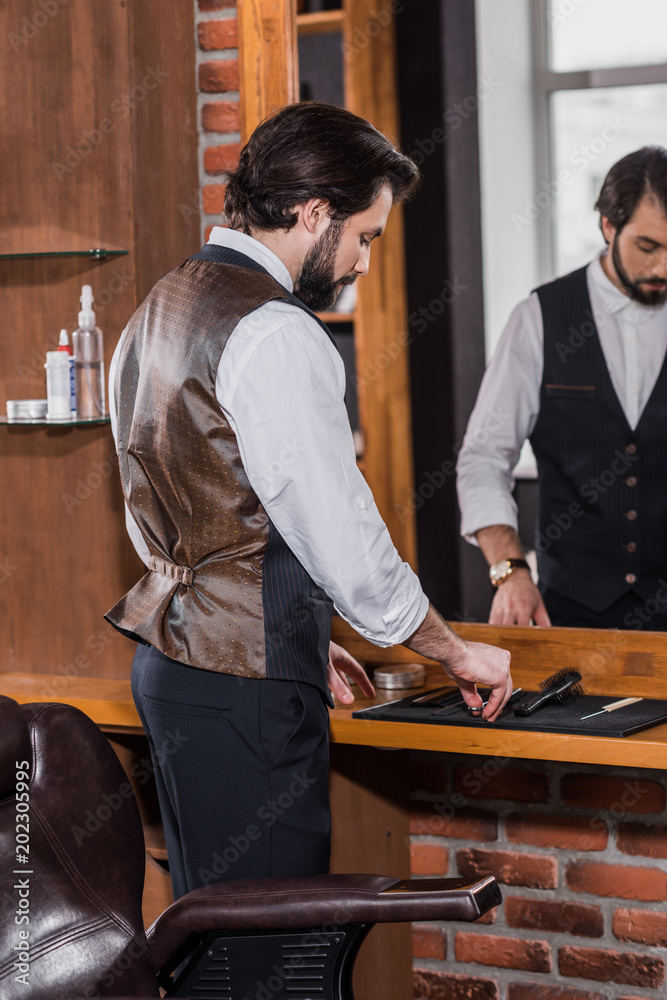 handsome young barber in stylish vest taking tools from rubber mat on workplace