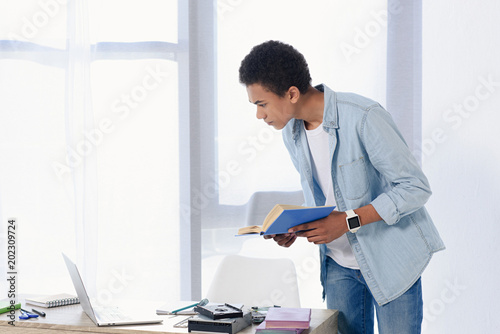 african american teenager holding book and looking at laptop at home