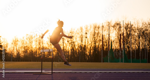 Athletic woman jumping above the hurdle on stadium running track during sunset